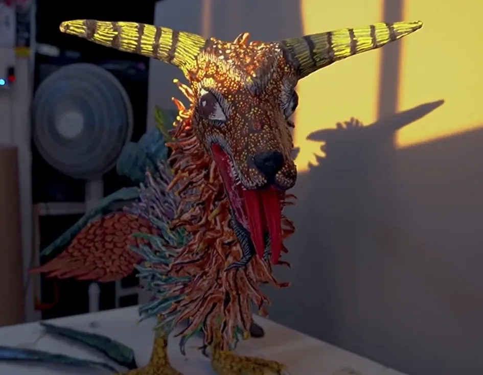 Alebrije with a goat head and striped horns, against the light in the workshop; its shadow falls on the wall.