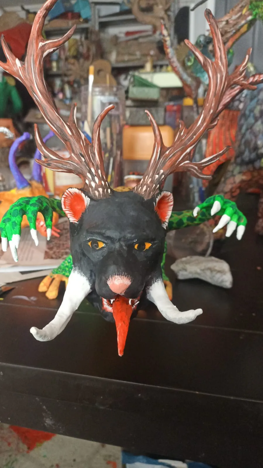 Black-headed, antlered alebrije on the workshop table, with paint jars and brushes behind.