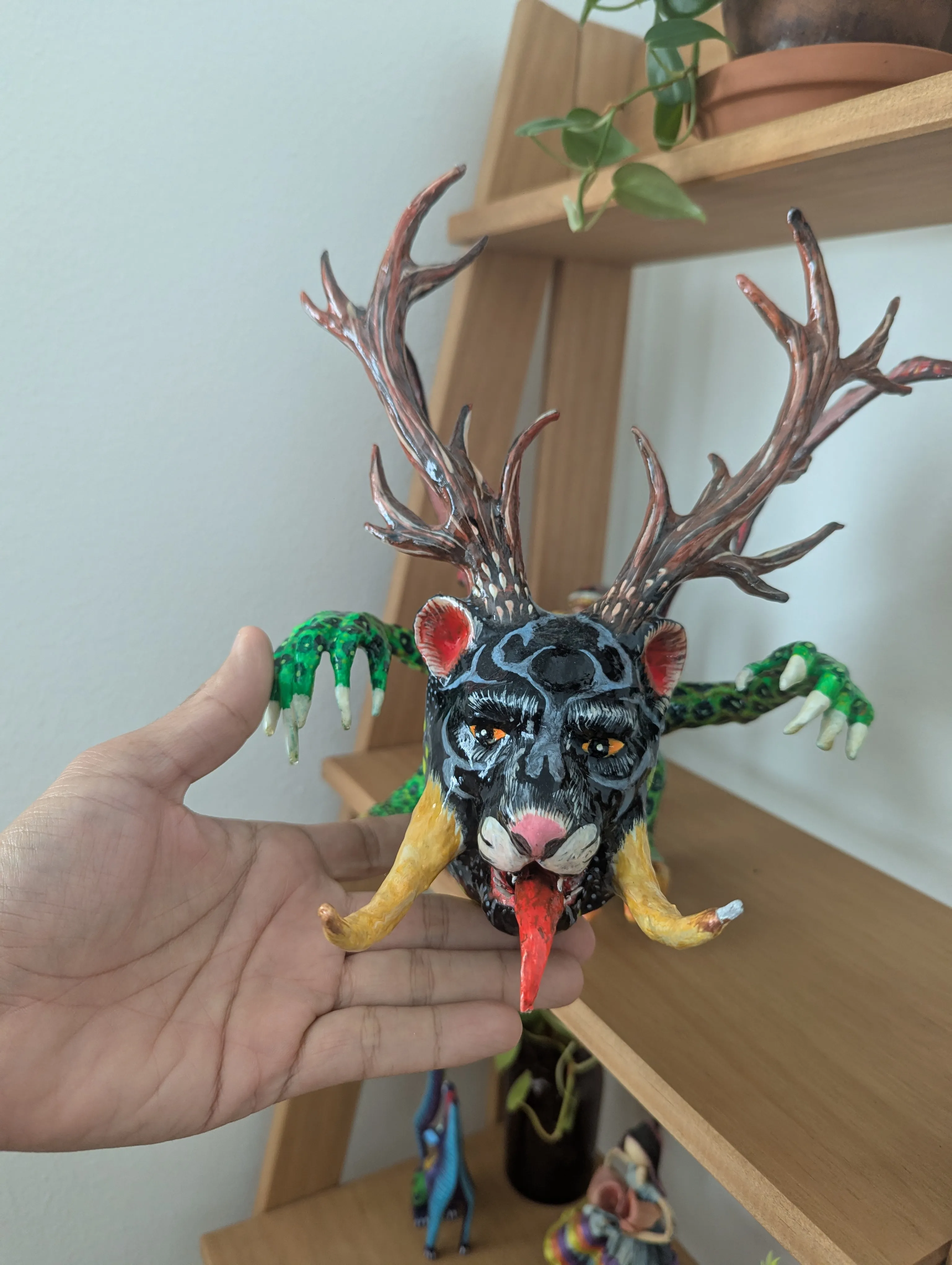 Una mano sostiene al alebrije de frente; se aprecian los ojos ámbar, el hocico rosado, los colmillos amarillos y la lengua roja.
