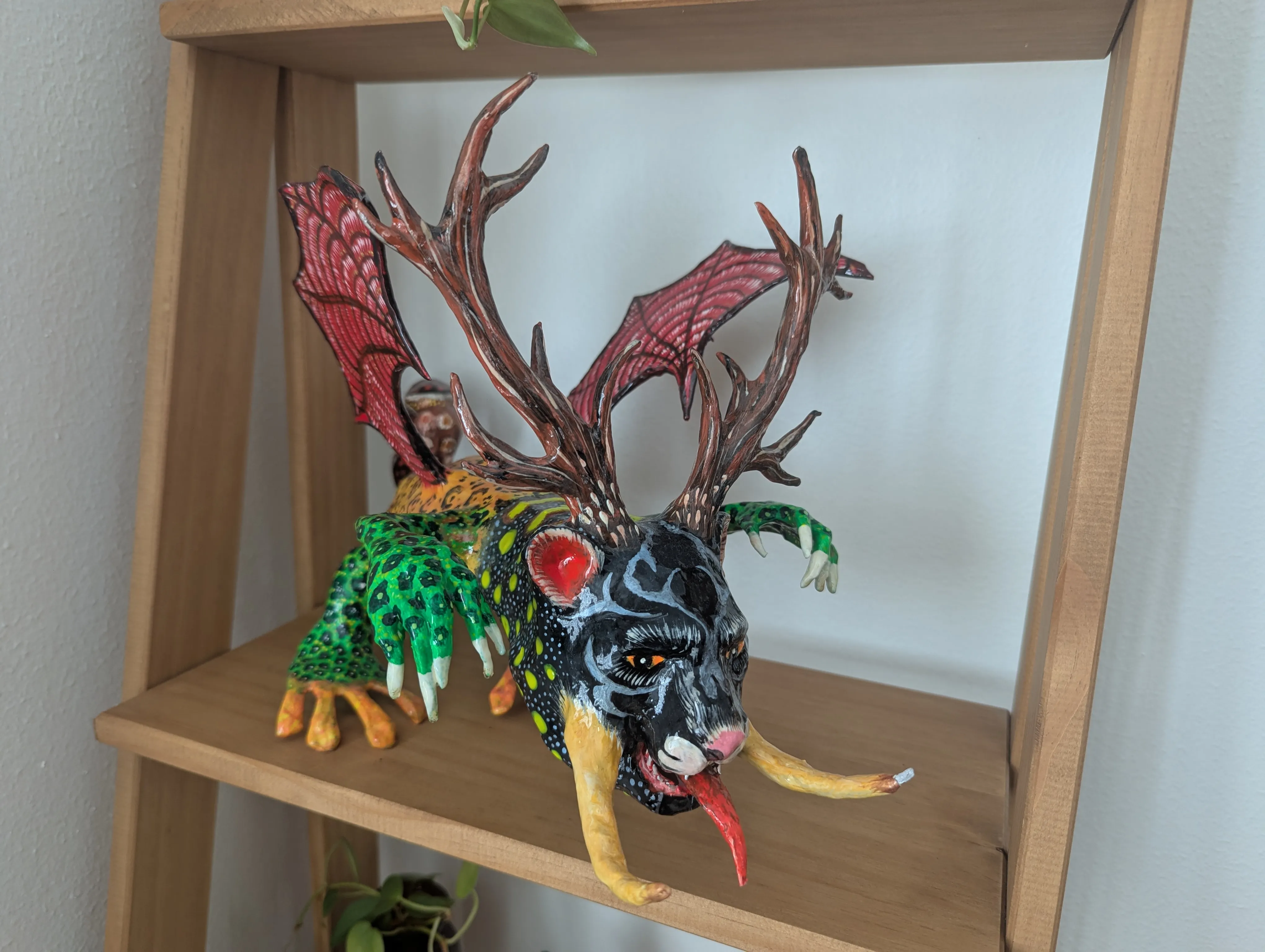 Cartonería alebrije with a black feline head, deer antlers, red bat wings and green claws, seen in three-quarter view on a shelf.