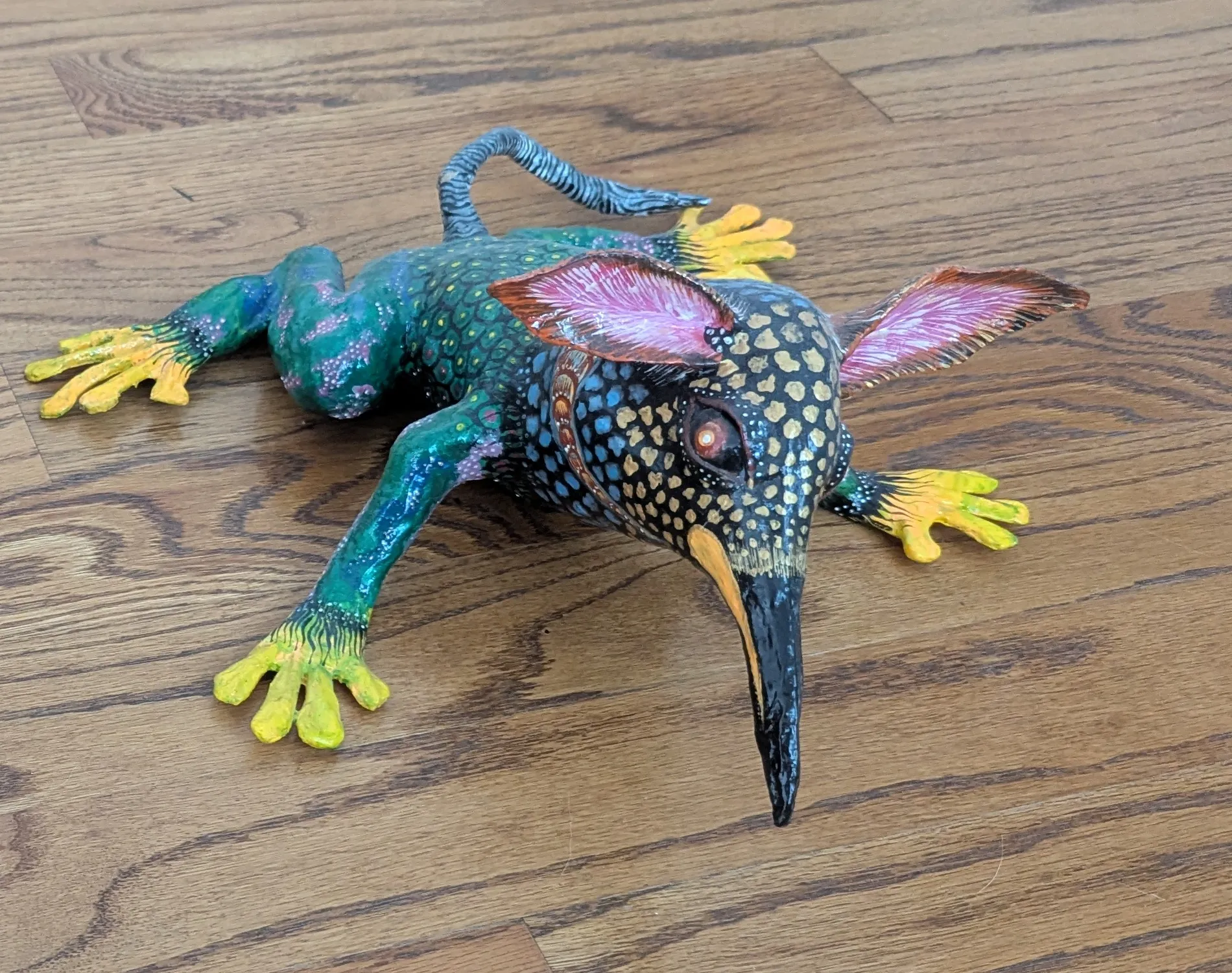 Alebrije with a green frog body, yellow legs, a bird head with a black beak and large pink ears, seen from the front.