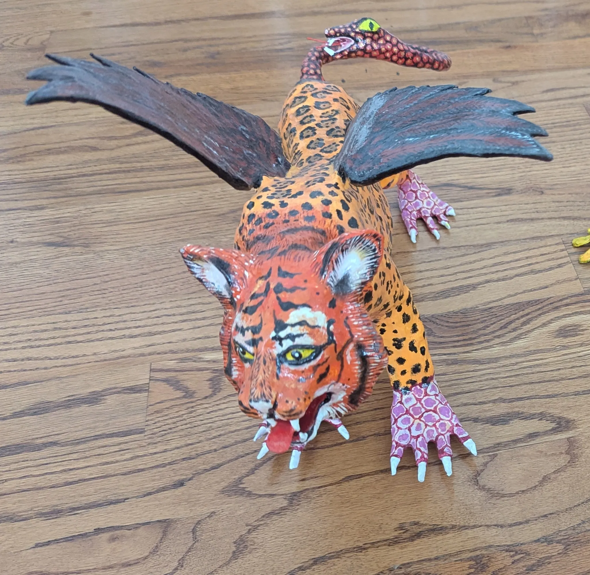 Alebrije with a roaring tiger head, jaguar-spotted body, spread black wings and pink scaled legs.