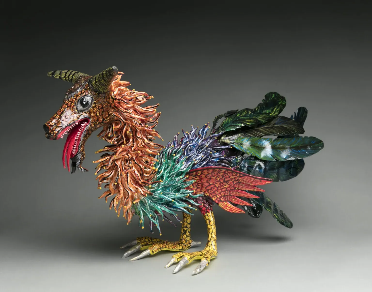 Cartonería alebrije with a head with striped horns, a large eye and a red tongue, a mane of orange strips, a rooster body with green, blue and purple feathers, a red wing and yellow legs, on a gray studio background.