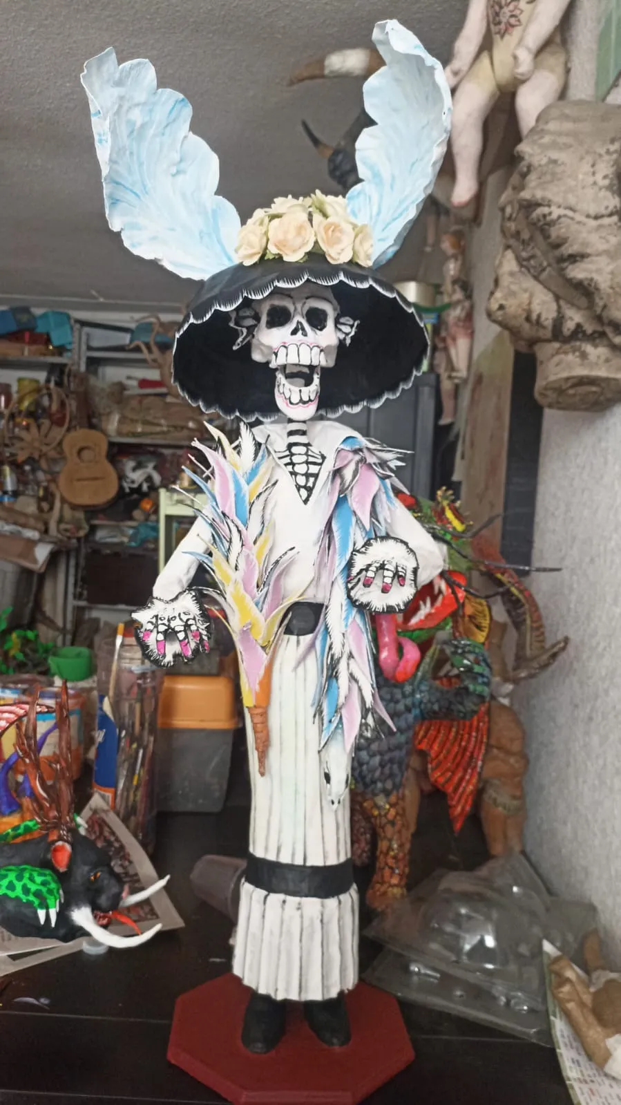 Cartonería Catrina with a black hat crowned with roses, two large blue plumes, a multicolored feather shawl and a white dress, standing on a red base in the workshop.