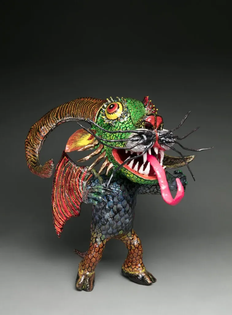 Cartonería alebrije with a scaly green head, a yellow eye, black whiskers, fangs and a pink tongue, a large curved striped horn, red fins, a body of blue scales and hoofed legs, on a gray studio background.