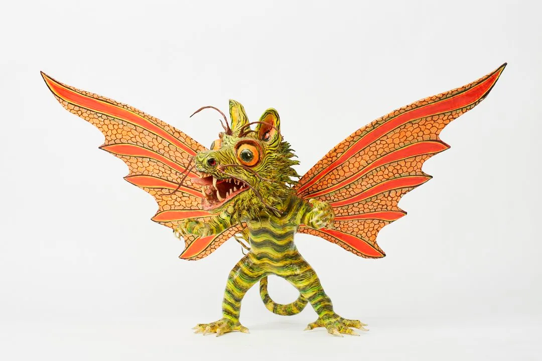 Cartonería alebrije with a green-and-yellow striped body, open orange-and-red wings and a fanged feline face.