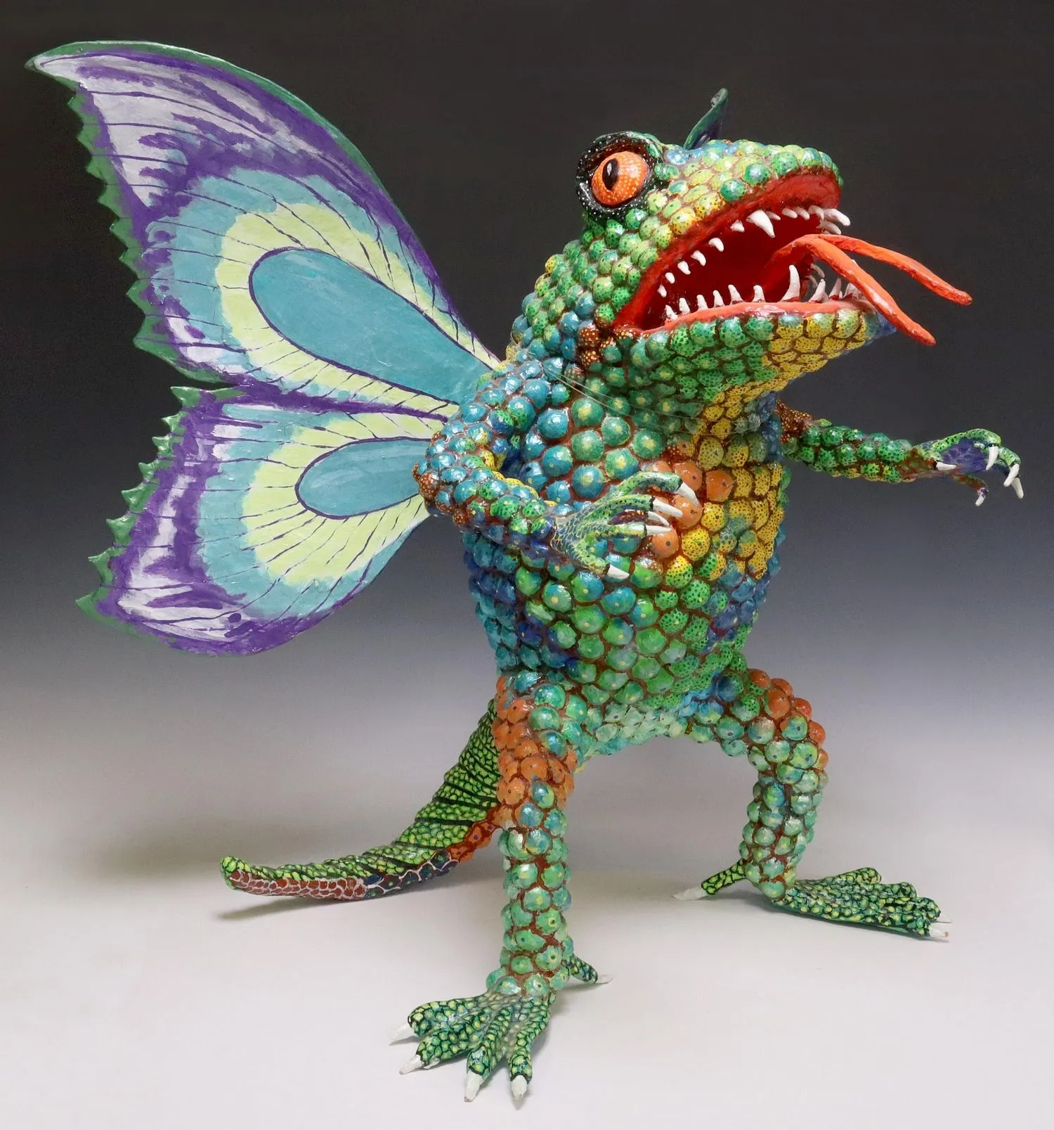 Alebrije with a frog body scaled in green, blue and orange, purple-and-blue butterfly wings and an open fanged mouth.
