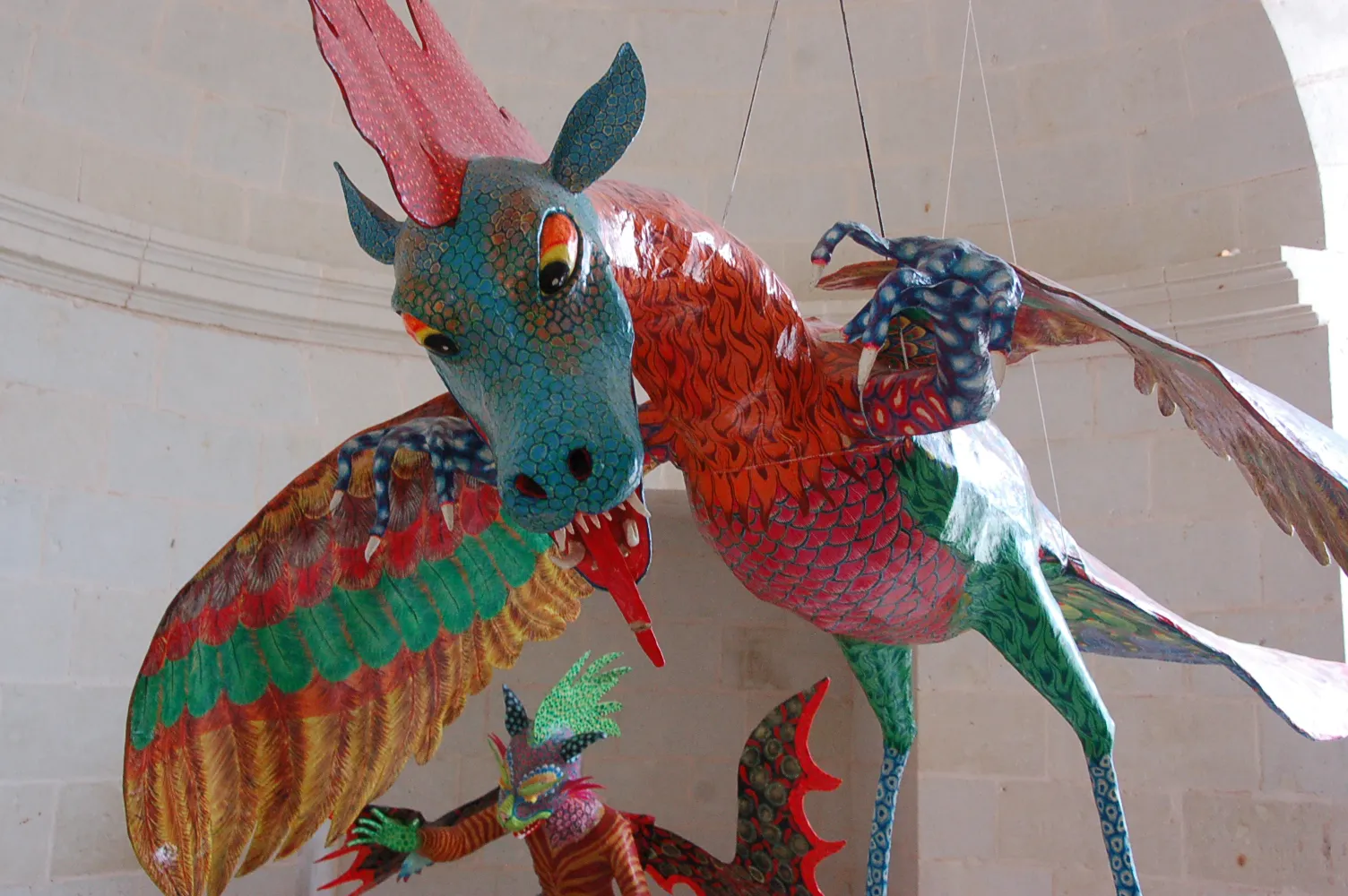 Cartonería dragon hung on wires in a pale stone room, with a blue-green head, red crest and tongue, scaly red body, wings of red, green and yellow feathers and long legs; the head of another alebrije peeks out below.