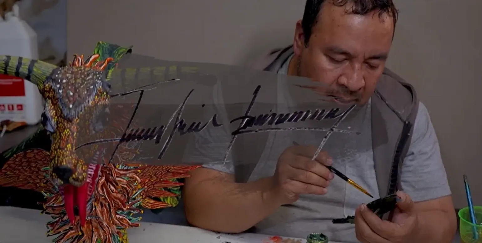 Felipe Linares Vargas paints with a fine brush at the workshop table; beside him, a horned alebrije with a multicolored mane.