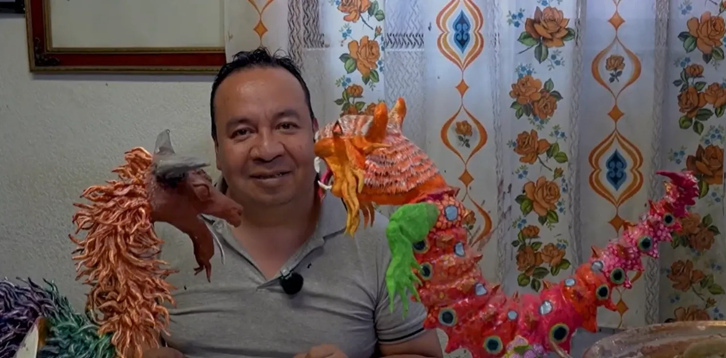 Felipe Linares Vargas smiles seated in his workshop, between two cartonería alebrijes.