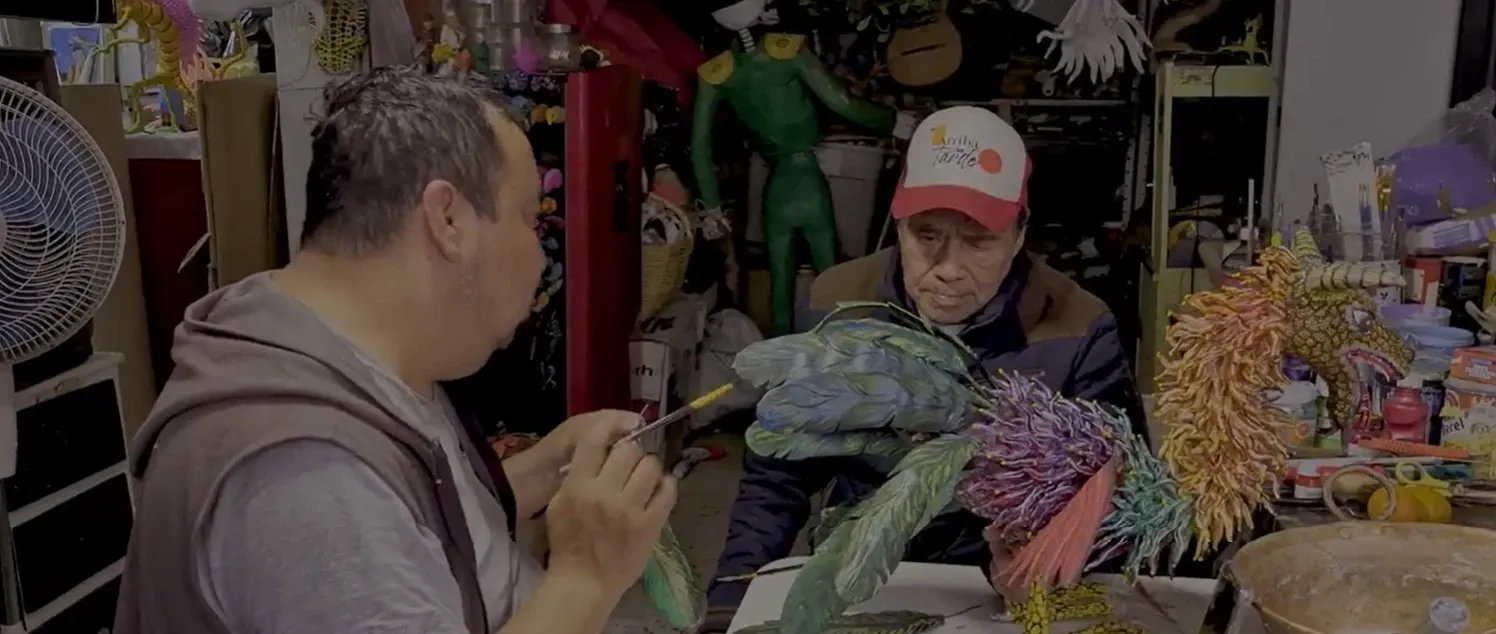 Felipe Linares Vargas paints an alebrije at the workshop table; across from him, his father Felipe Linares Mendoza watches the piece.
