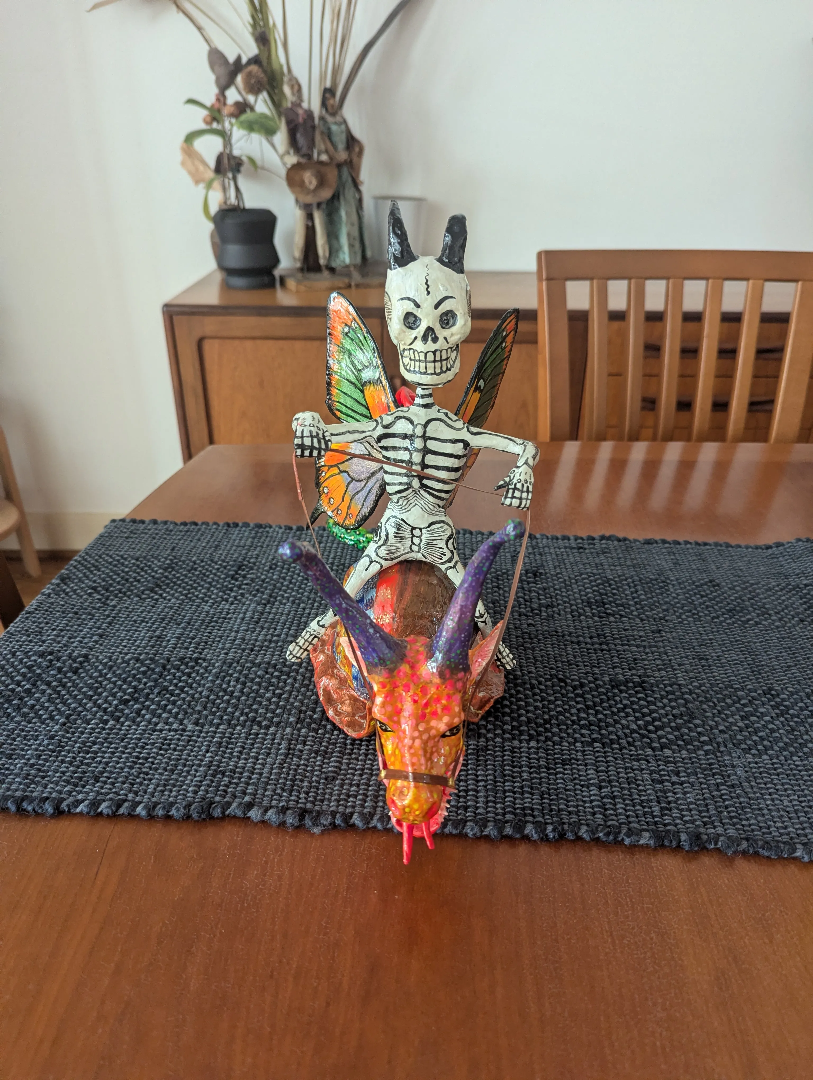 Cartonería skeleton with horns and monarch-butterfly wings, riding an orange creature with purple horns and wire reins, on a table.