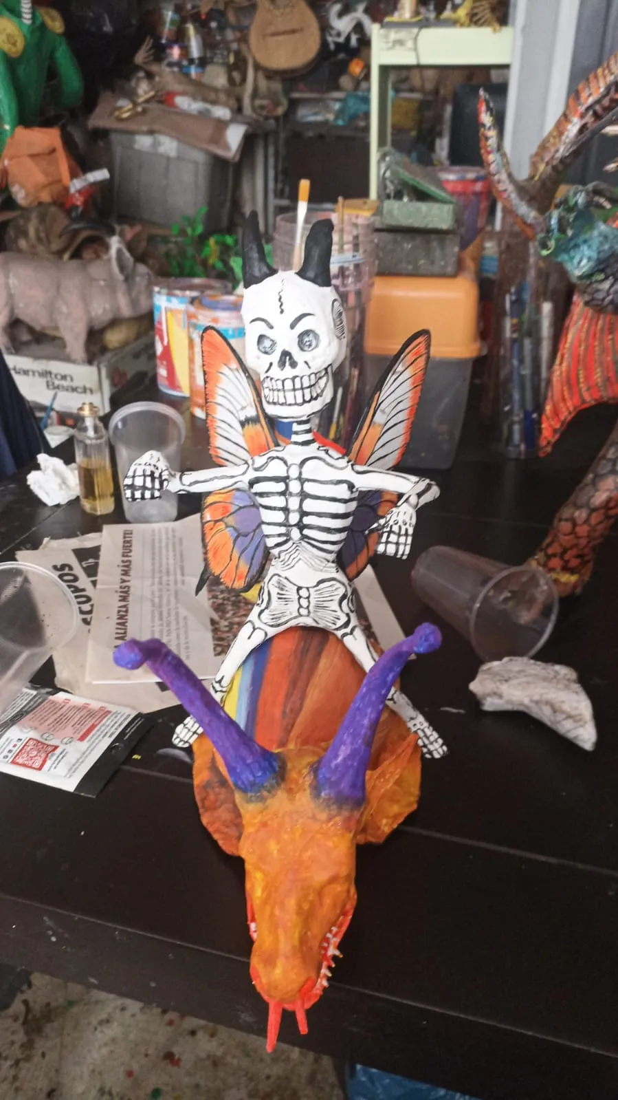 Skeleton with butterfly wings riding an orange creature, in progress among cups and brushes.