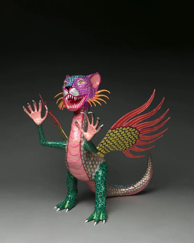 Cartonería alebrije: a standing winged lion with a purple and pink head, fangs, orange whiskers, raised pink hands, a green and pink body, yellow and red wings and a long tail, on a gray studio background.