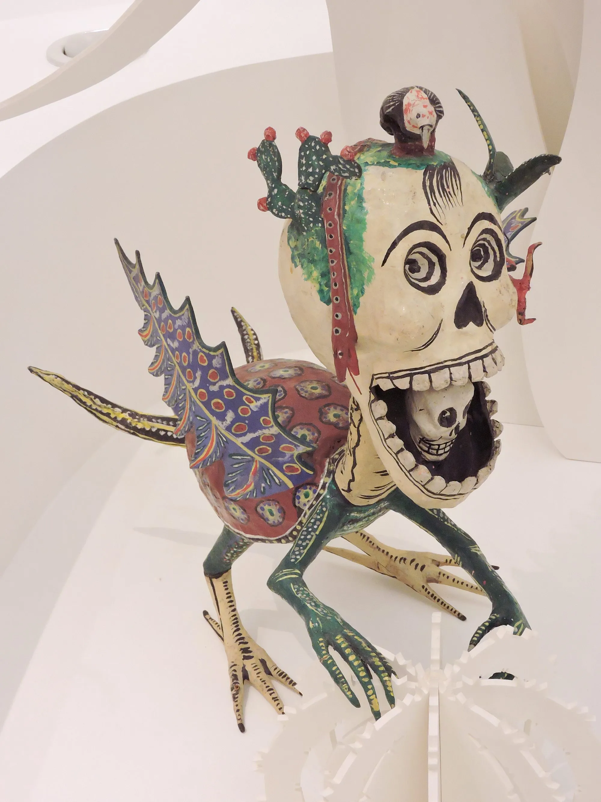 Alebrije with a white skull for a head and an open mouth from which a smaller skull peeks out; on top of the head, prickly pears with red fruit and a dark bird; a red body with flowers, jagged blue wings, green front legs and cream hind legs, clawed, on a white stand.