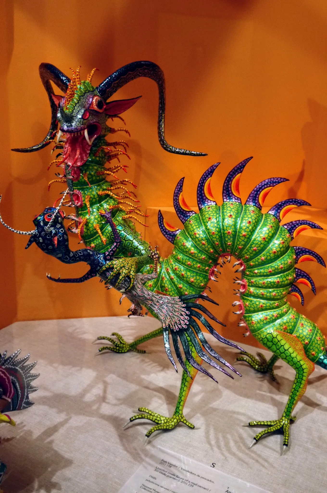 Cartonería dragon with a long, arched green body, segmented and dotted in red, purple and orange crests along its back, black ram’s horns, an open fanged mouth and green clawed legs, against an orange background.