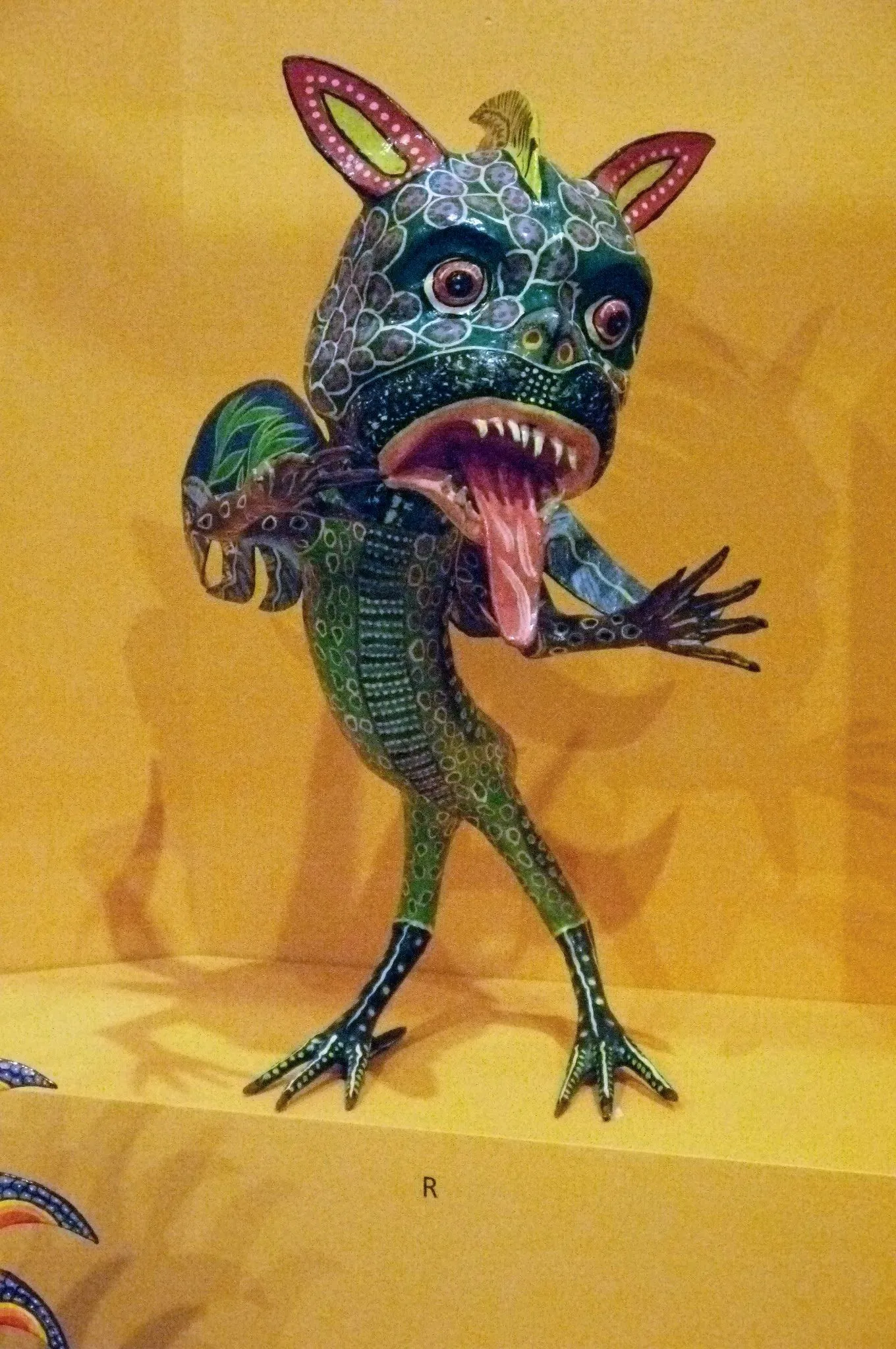 Green alebrije standing on long bird’s legs, with a large scaled head, pointed pink ears, bulging eyes, fangs and a long pink tongue hanging out; a small wing on its back, against a yellow background.