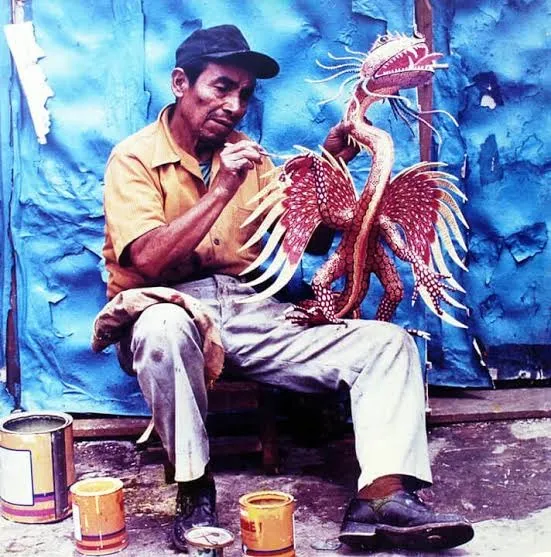Fotografía antigua: Pedro Linares López, con gorra y camisa amarilla, sentado ante un muro azul, pinta con un pincel un alebrije rojo con alas y cresta; latas de pintura en el suelo.